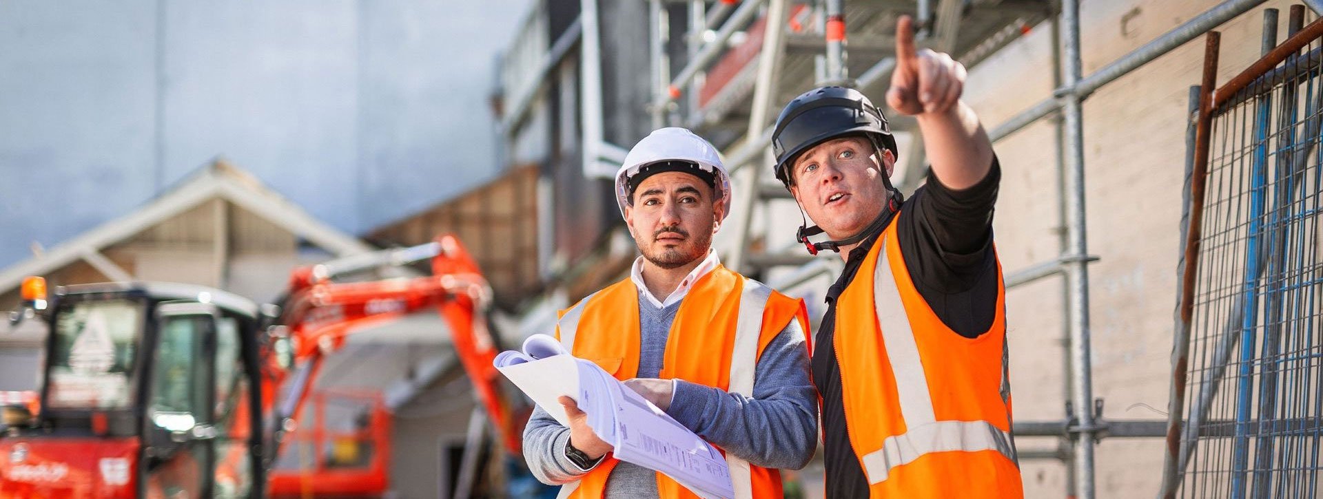 Construction site scene with two workers in safety gear, one pointing while holding plans